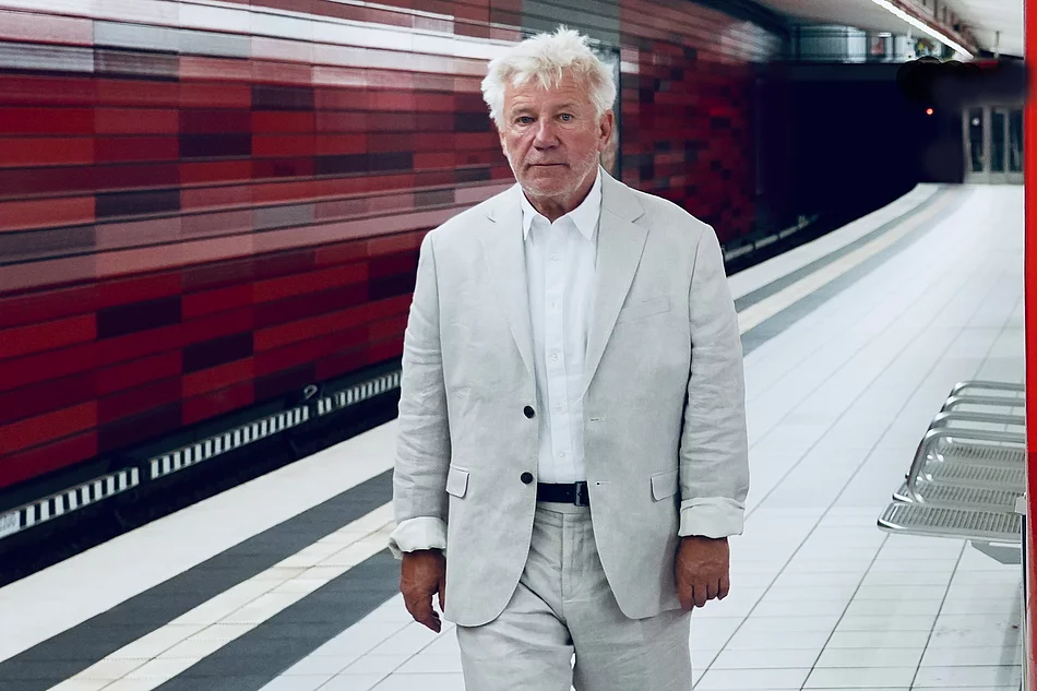 Schauspieler Jörg Schüttauf geht in einer U-Bahnstation entlang. Er trägt einen hellen Anzug. Die Wand hinter ihm ist rot.