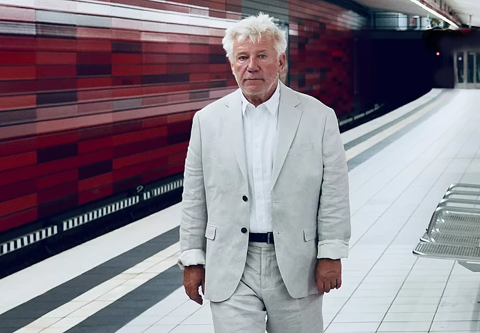 Schauspieler Jörg Schüttauf geht in einer U-Bahnstation entlang. Er trägt einen hellen Anzug. Die Wand hinter ihm ist rot.