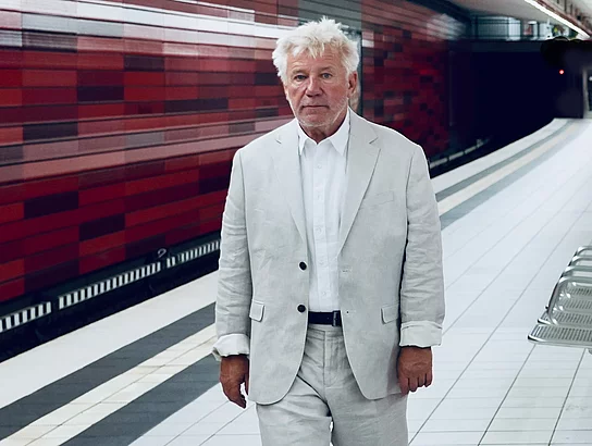 Schauspieler Jörg Schüttauf geht in einer U-Bahnstation entlang. Er trägt einen hellen Anzug. Die Wand hinter ihm ist rot.