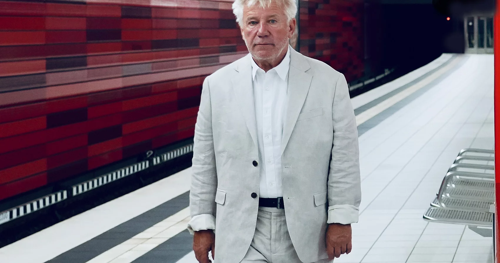 Schauspieler Jörg Schüttauf geht in einer U-Bahnstation entlang. Er trägt einen hellen Anzug. Die Wand hinter ihm ist rot.