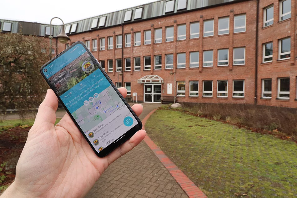 Eine Hand hält ein Smartphone mit der PPush-App vor dem Glinder Rathaus 