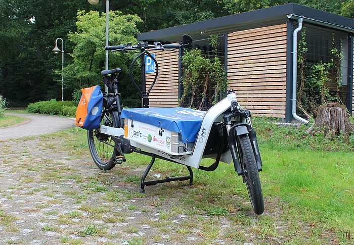 Ein Fahrrad mit einem großen flachen Behälter hinter dem Vorderrad für Lasten. 