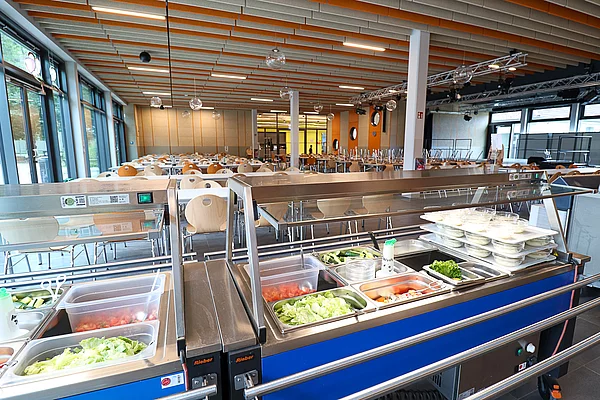Im Vordergrund eine Salatbar, im Hintergrund Stühle einer Mensa.