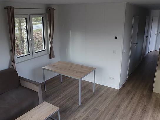 In einem Raum steht ein Tisch und ein Sofa. Rechts ist eine Küchenzeile im Flur zu erkennen.