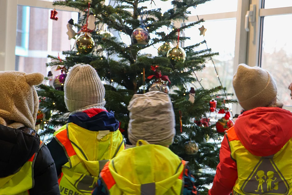 Kinder mit Jacken und Mütze stehen vor einem Weihnachtsbaum.