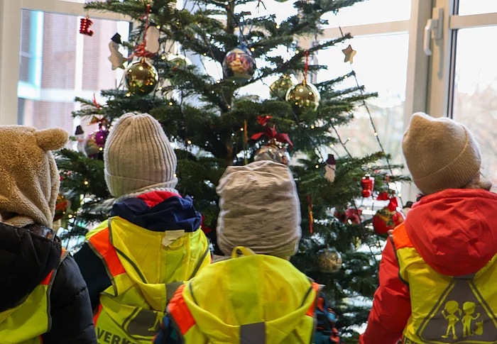 Kinder mit Jacken und Mütze stehen vor einem Weihnachtsbaum.