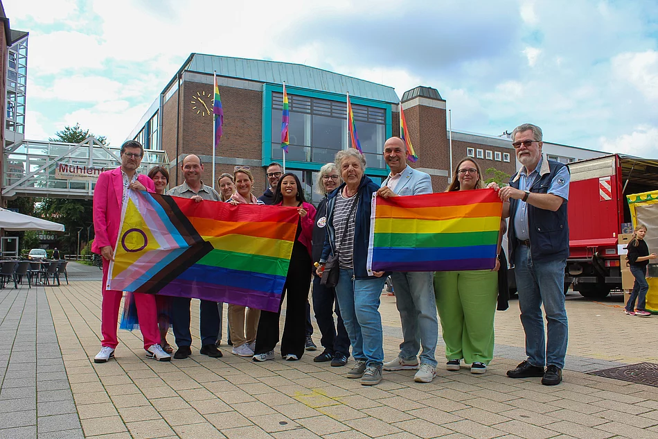 Menschen mit Regenbogenflaggen stehen vor dem Bürgerhaus. An den Fahnenmasten sind ebenfalls Regenbogenfahnen zu sehen.