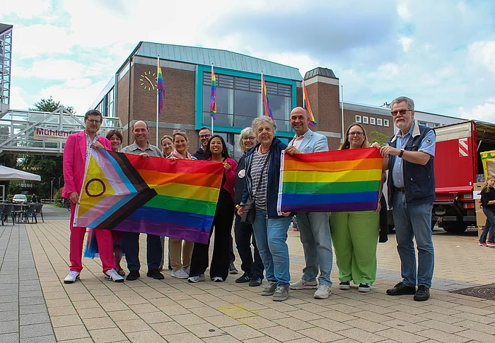 Menschen mit Regenbogenflaggen stehen vor dem Bürgerhaus. An den Fahnenmasten sind ebenfalls Regenbogenfahnen zu sehen.