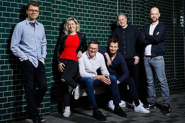 Fünf Männer und eine Frau stehen zusammen an einer gefliesten Wand für ein Bandfoto.