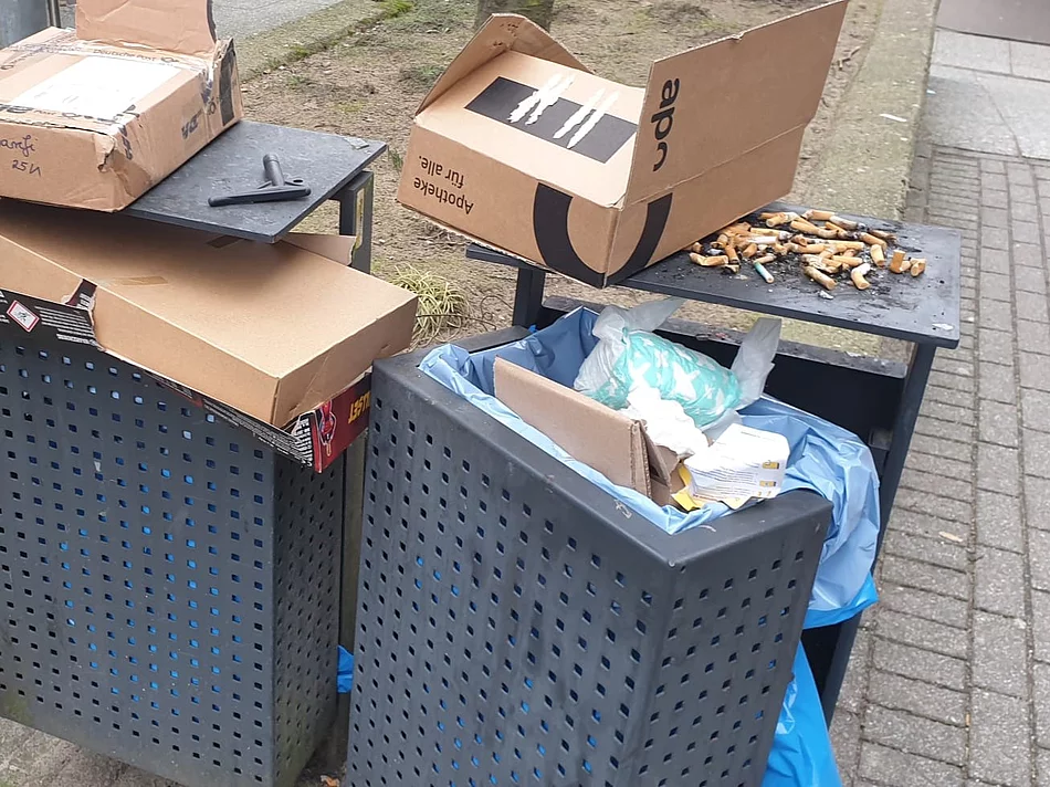 Zwei graue Mülleimer sind vollgestopft mit Paketkartons. Auf dem Mülleimer liegen sehr viele Zigarettenstummel. 