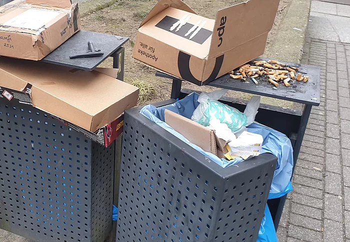 Zwei graue Mülleimer sind vollgestopft mit Paketkartons. Auf dem Mülleimer liegen sehr viele Zigarettenstummel. 