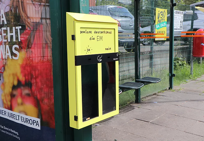 Eine schmale gelbe Box für Zigarettenstummel mit zwei Löchern. Jedes Loch führt in ein eigenes Fach. Dieser Abstimmungsaschenbecher hängt an einer Bushaltestelle.