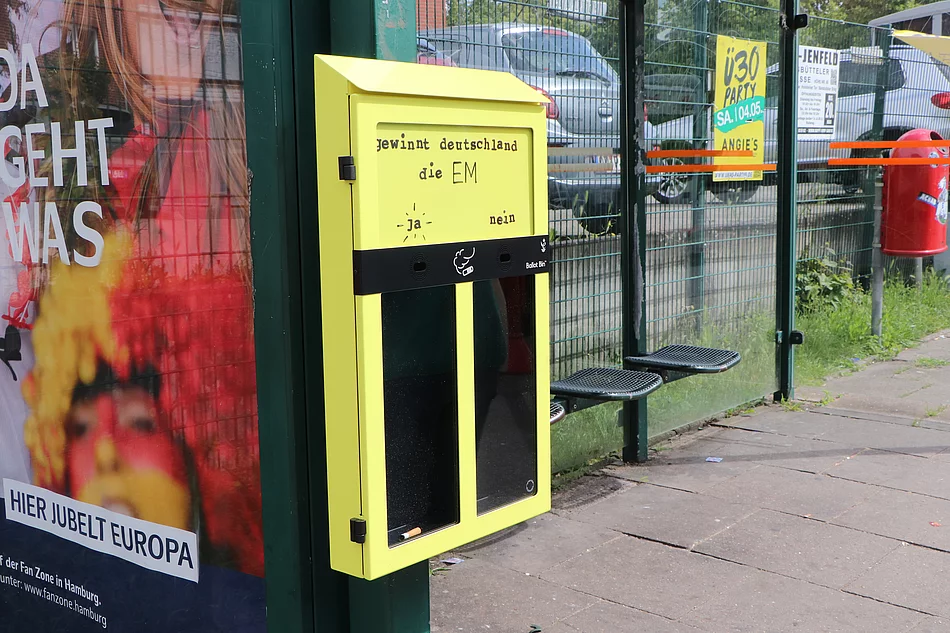Eine schmale gelbe Box für Zigarettenstummel mit zwei Löchern. Jedes Loch führt in ein eigenes Fach. Dieser Abstimmungsaschenbecher hängt an einer Bushaltestelle.