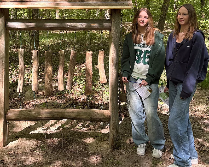 Zwei Mädchen stehen neben senkrecht aufgehängten Holzstämmen, einem Dendrophon. 