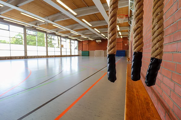 Blick in eine Turnhalle, links bodentiefe Fenster, rechts hängen drei Kletterseile