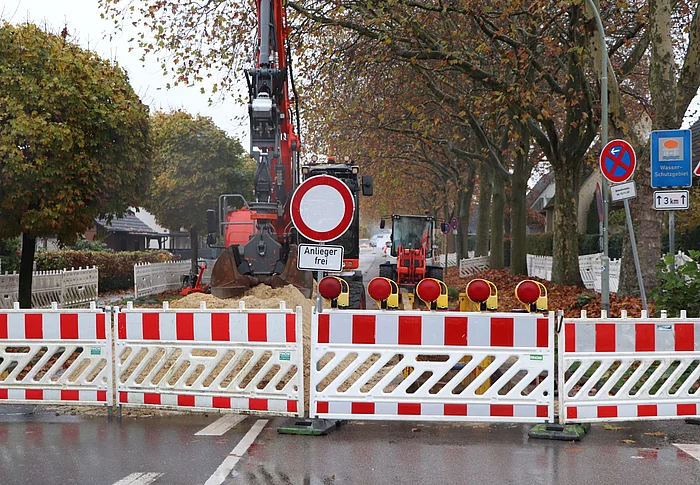 Eine Straße ist mit rot-weissen Absperrelementen blockiert. Dahinter ein Bagger und Baumaterial.