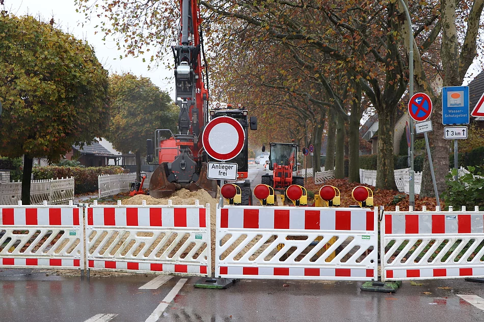Eine Straße ist mit rot-weissen Absperrelementen blockiert. Dahinter ein Bagger und Baumaterial.