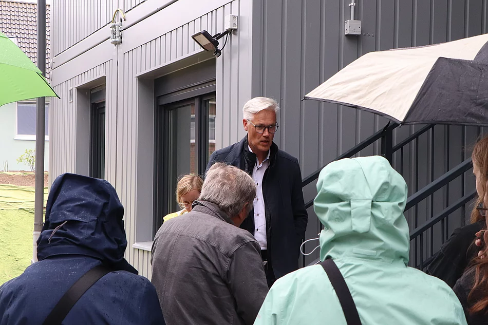 Bürgermeister Rainhard Zug spricht zu Menschen mit Regenjacken und Schirmen vor einem grauen Haus.