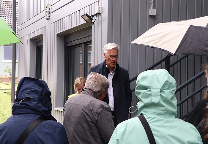 Bürgermeister Rainhard Zug spricht zu Menschen mit Regenjacken und Schirmen vor einem grauen Haus.