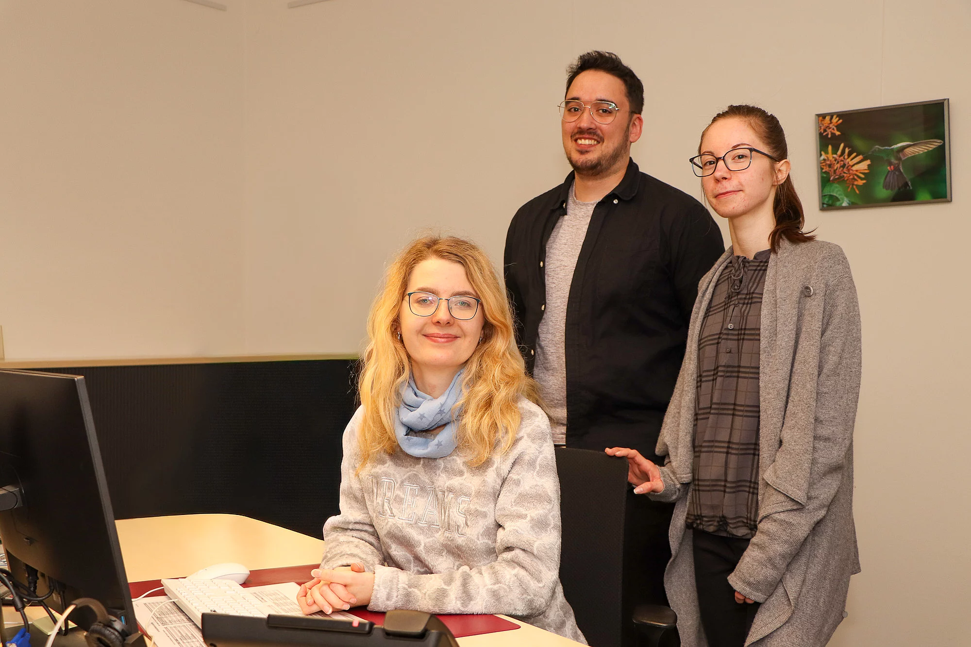 Zwei junge Frauen und ein junger Mann stehen an einem Schreibtisch. 