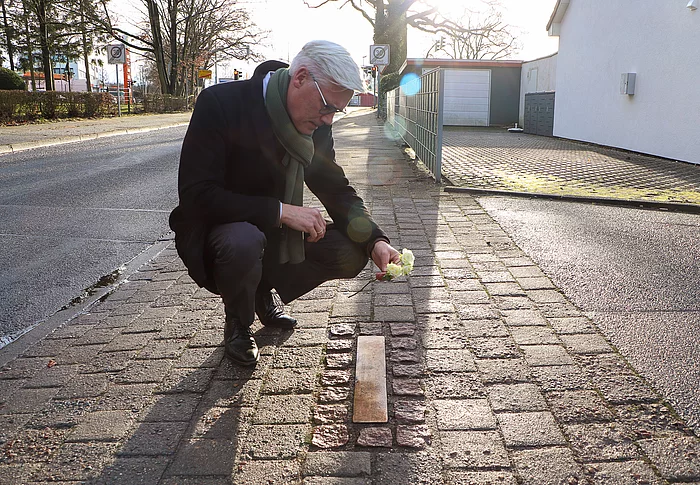 Glindes Bürgermeister Rainhard Zug kniet mit einer weißen Rose an der Stolperschwelle für Zwangsarbeitslager Wiesenfeld.