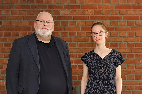 Ein Mann mit weißem Bart und eine junge Frau stehen vor einer Backsteinwand.