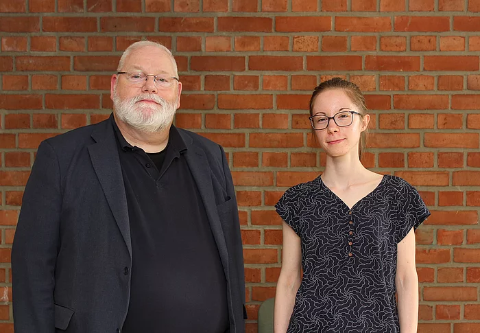 Ein Mann mit weißem Bart und eine junge Frau stehen vor einer Backsteinwand.