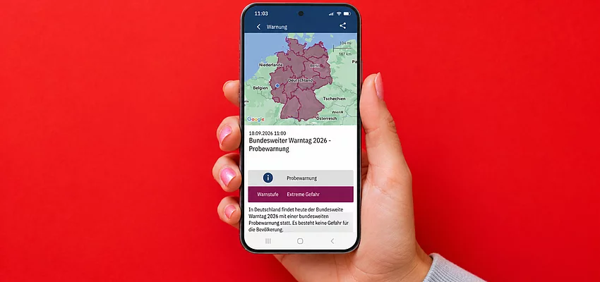 Warntag Hinweis Handy Eine Hand hält ein Handy mit einer Meldung zum Warntag. Der Hintergrund ist rot.
