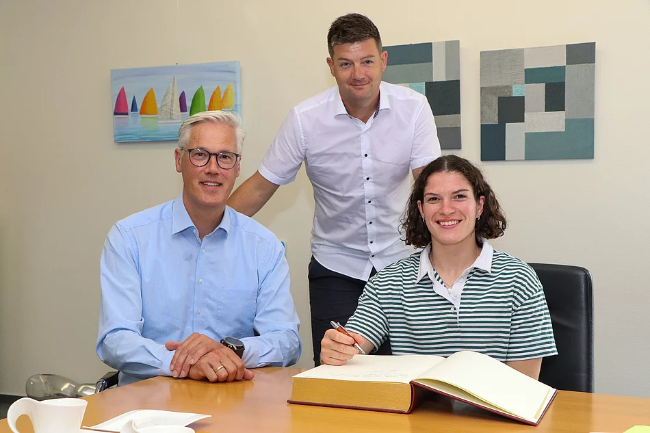 Seija Ballhaus sitzt am goldenen Buch und Bürgermeister Rainhard Zug und TSV Geschäftsführer Carsten Henning sind neben ihr. 