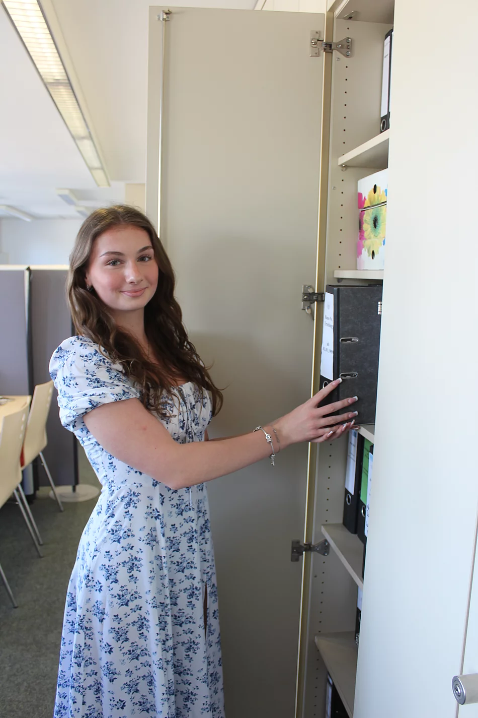 Eine junge Frau in einem Blümchenkleid steht an einem Schrank mit einem Aktenordner