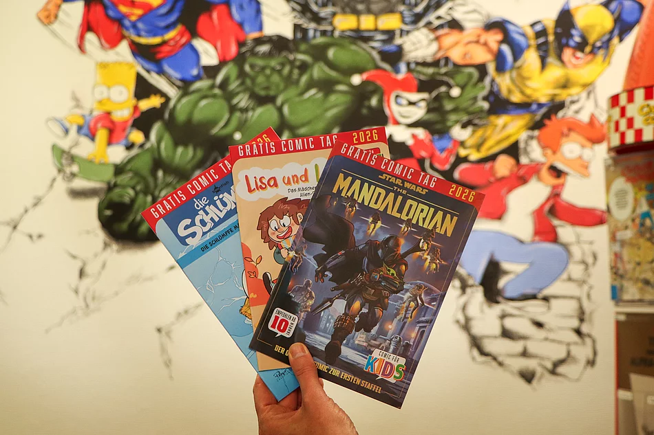 Vor einer Wand mit Superheldenzeichnungen werden drei Comichefte präsentiert.