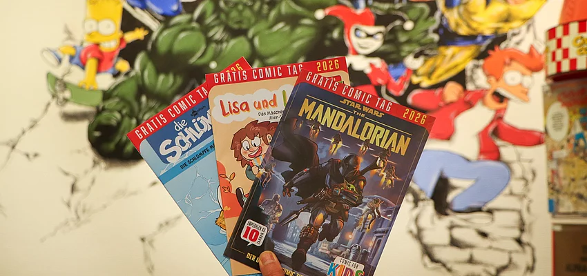 Vor einer Wand mit Superheldenzeichnungen werden drei Comichefte präsentiert.