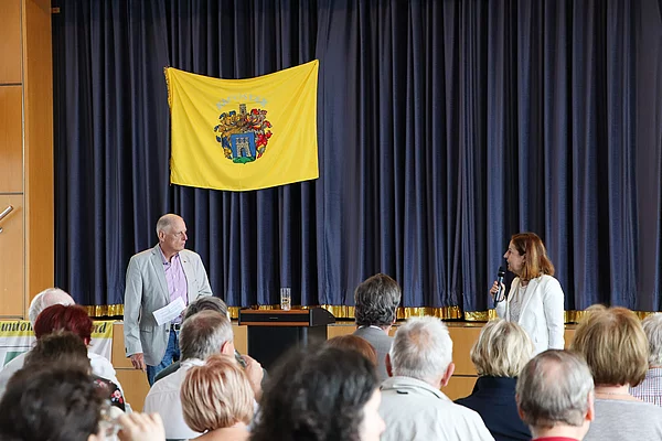 Ein Mann und eine Frau sprechen zum Publikum. Im Hintergrund ist das Wappen von Kaposvar an Bühnenvorhang angebracht.