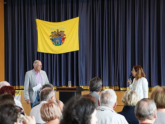 Ein Mann und eine Frau sprechen zum Publikum. Im Hintergrund ist das Wappen von Kaposvar an Bühnenvorhang angebracht.