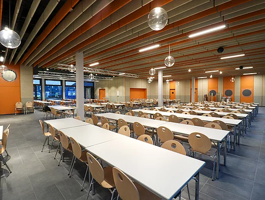In der Mensa stehen viele Tische und Stühle in Reihe. An der Decke hängen orangefarbene Platten als Schallschutzelemente.