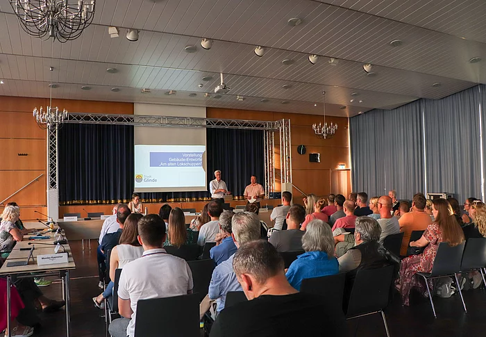 Publikum schaut zu einer Leinwand vor der Bürgermeister Rainhard Zug steht.
