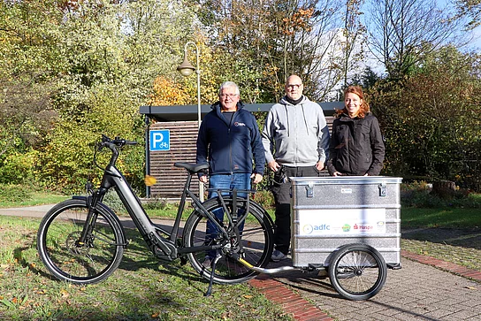 zwei Männer und eine Frau stehen an einem E-Bike mit einem silbernen Anhänger.