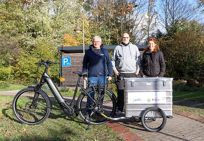 zwei Männer und eine Frau stehen an einem E-Bike mit einem silbernen Anhänger.