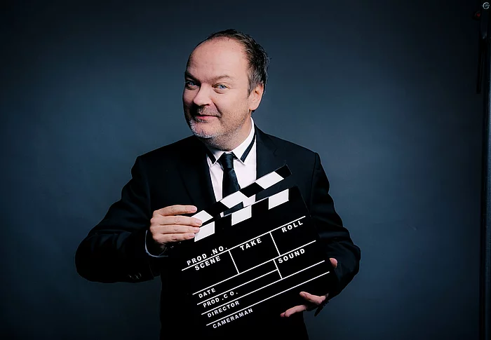 Ein Mann mit Halbglatze hat eine Filmklappe vor sich.