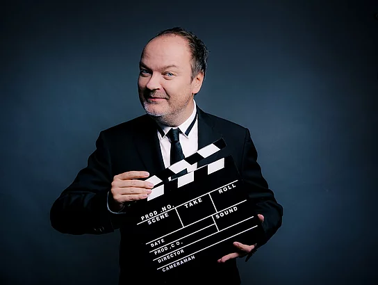 Ein Mann mit Halbglatze hat eine Filmklappe vor sich.