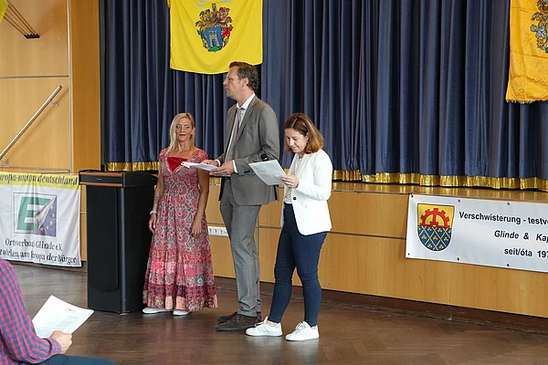 Zwischen zwei Frauen steht ein großer Mann der zum Publikum spricht. 