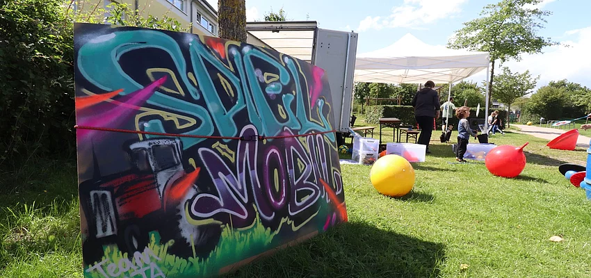 Kinder spielen auf einer Wiese, dafür ein Schild mit der Aufschrift "Spielmobil"