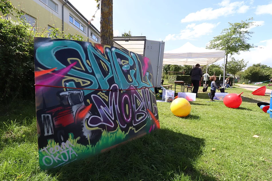 Kinder spielen auf einer Wiese, dafür ein Schild mit der Aufschrift "Spielmobil"