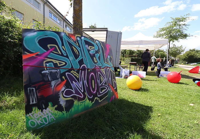 Kinder spielen auf einer Wiese, dafür ein Schild mit der Aufschrift "Spielmobil"