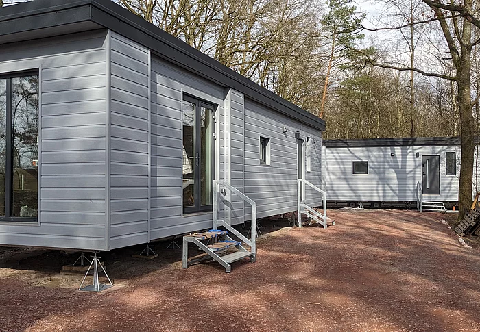 Die Außenansicht von zwei Mobilheimen für Geflüchtete. Sie haben ein schwarzes Flachdach und eine graue Fassade. Die Gebäude sind aufgebockt, wie ein Anhänger.