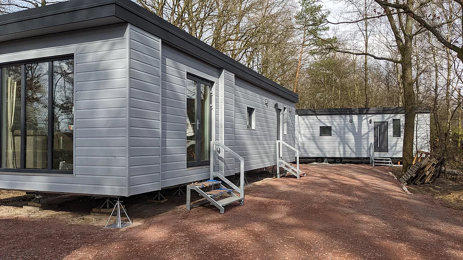 Die Außenansicht von zwei Mobilheimen für Geflüchtete. Sie haben ein schwarzes Flachdach und eine graue Fassade. Die Gebäude sind aufgebockt, wie ein Anhänger.