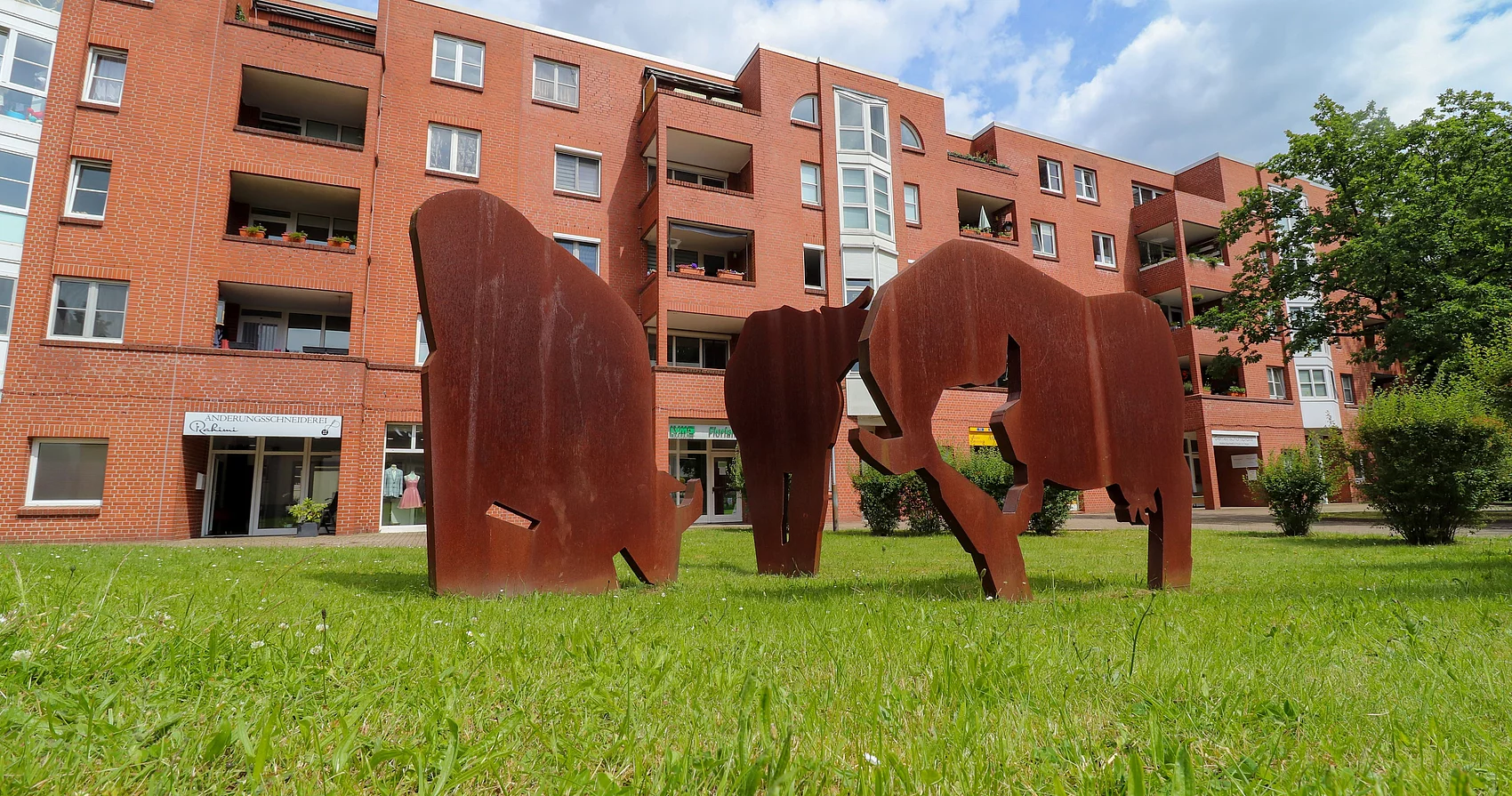 Drei Kühe aus rostigem Metall stehen auf einer Wiese vor einem Mehrfamilienhaus.