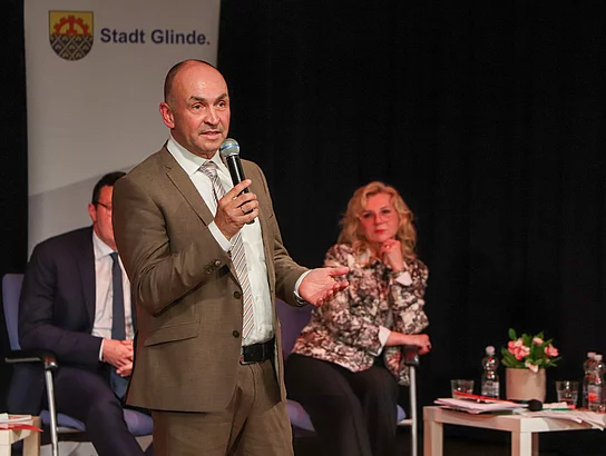 Oliver Sendzik steht auf einer Bühne und hat ein Mikrofon in der Hand.