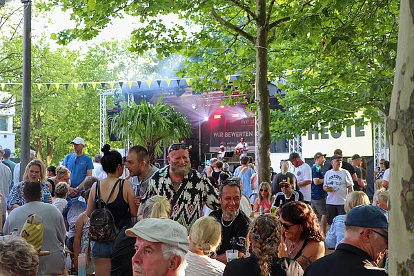 Unter Bäumen sitzen Menschen auf Bierzelt-Garnituren und schauen zur Bühne