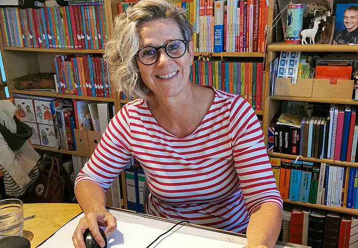 Eine Frau mit grauen Locken und schwarzer Brille lächelt freundlich in die Kamera.
